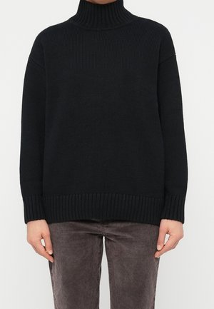 Persoon die een zwarte geribde coltrui en donkergrijze broek draagt, staat met ontspannen handen langs het lichaam.