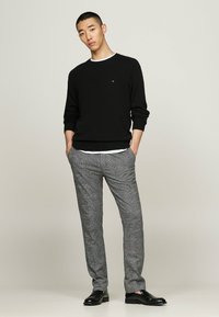 Junger Mann steht mit Händen in den Taschen, trägt einen schwarzen Pullover, graue Karohosen und schwarze Slipper vor einem schlichten Hintergrund.