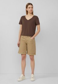 T-shirt marron à col en V associé à un short beige retroussé. Le mannequin porte des baskets blanches, mettant en avant une tenue décontractée et contemporaine.
