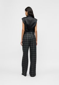 Γυναίκα με μακριά σκούρα μαλλιά, φορώντας αμάνικο μαύρο σατέν τοπ με δέσιμο στην πλάτη και ψηλόμεσα, καρό ασπρόμαυρα φαρδιά παντελόνια.