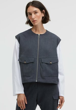 Graue ärmellose Jacke mit vorderem Reißverschluss, zwei Brusttaschen mit Knöpfen und cropped Design, über einem weißen Langarmhemd getragen.