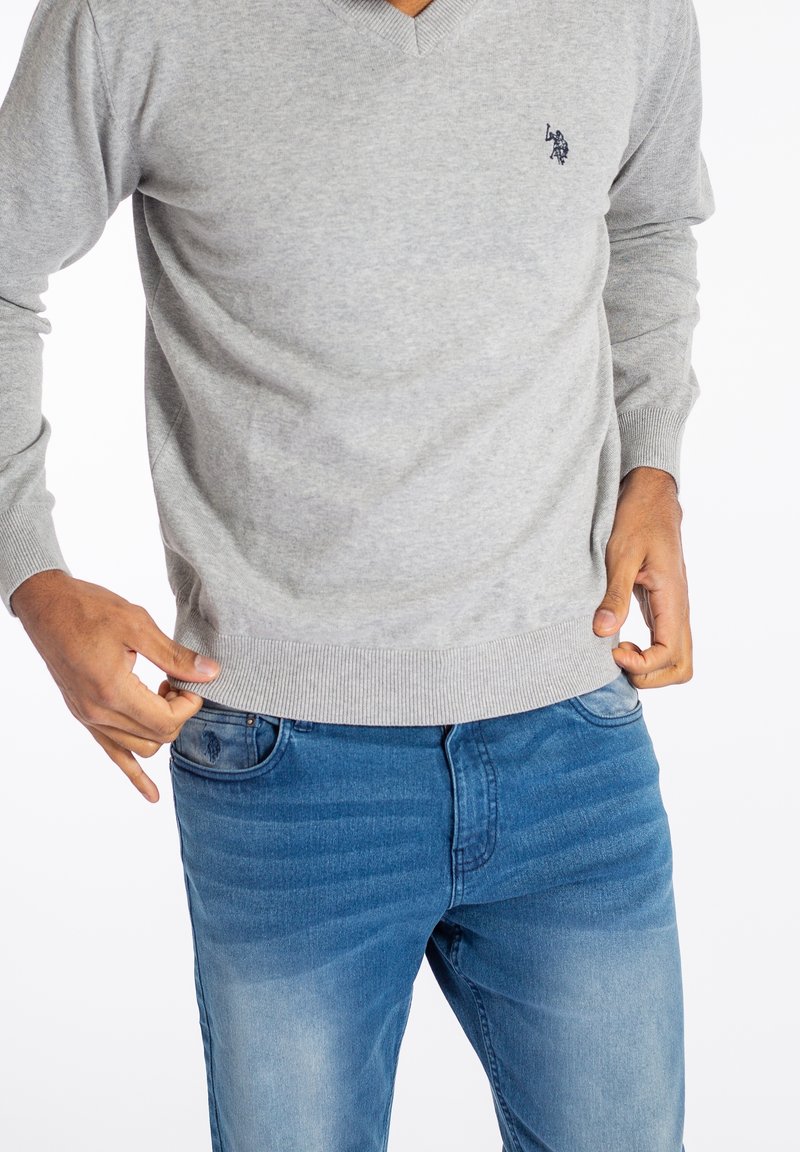Homme portant un pull gris clair à col en V et un jean bleu, ajustant l'ourlet du pull avec ses deux mains.
