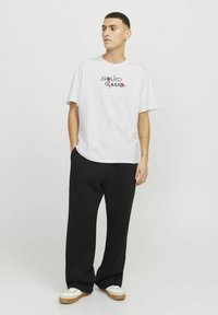Hvid bomulds T-shirt med "SQUID GAME" trykt i multicolor tekst på brystet. Kombineret med løse sorte bukser og hvide sneakers.
