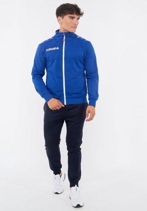 Homme portant une veste bleue zippée avec le texte "LEGEA", un pantalon de jogging noir et des baskets blanches, debout et regardant sur le côté sur un fond blanc.