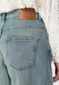 Jeans de ganga azul claro com um remendo de couro castanho rotulado "NOISY". Apresenta um bolso traseiro com detalhes sutis de costura.