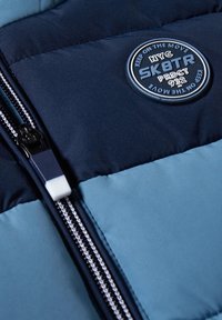 Veste matelassée bleue et marine avec une fermeture éclair et un écusson rond portant l'inscription « NYC SK8TR PRDCT 932 Keep On The Move » sur la poitrine.