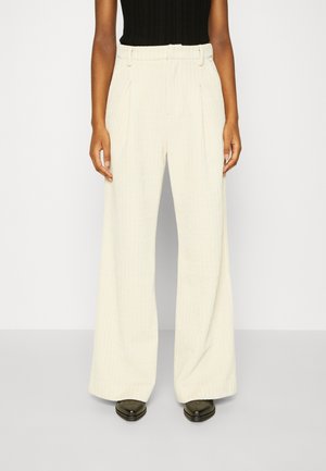 Kobieta ubrana w kremowe, prążkowane spodnie z wysokim stanem i szerokimi nogawkami z przednimi zakładkami oraz oliwkowozielone buty, stojąca na tle gładkiej białej ściany.