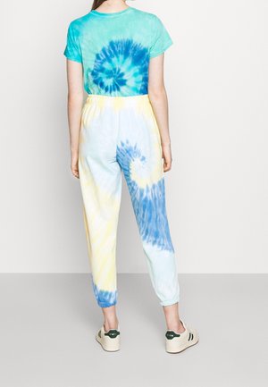 Henkilö, jolla on päällään sinikeltainen liukuvärjättyä jogger-housut ja vastaava turkoosi liukuvärjätty t-paita, seisoo yksiväristä taustaa vasten.