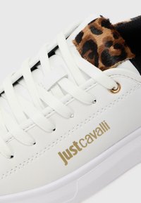 Bijele tenisice s bijelim vezicama, Just Cavalli logotip u zlatnoj boji i detalj leoparda blizu ovratnika oko gležnja.