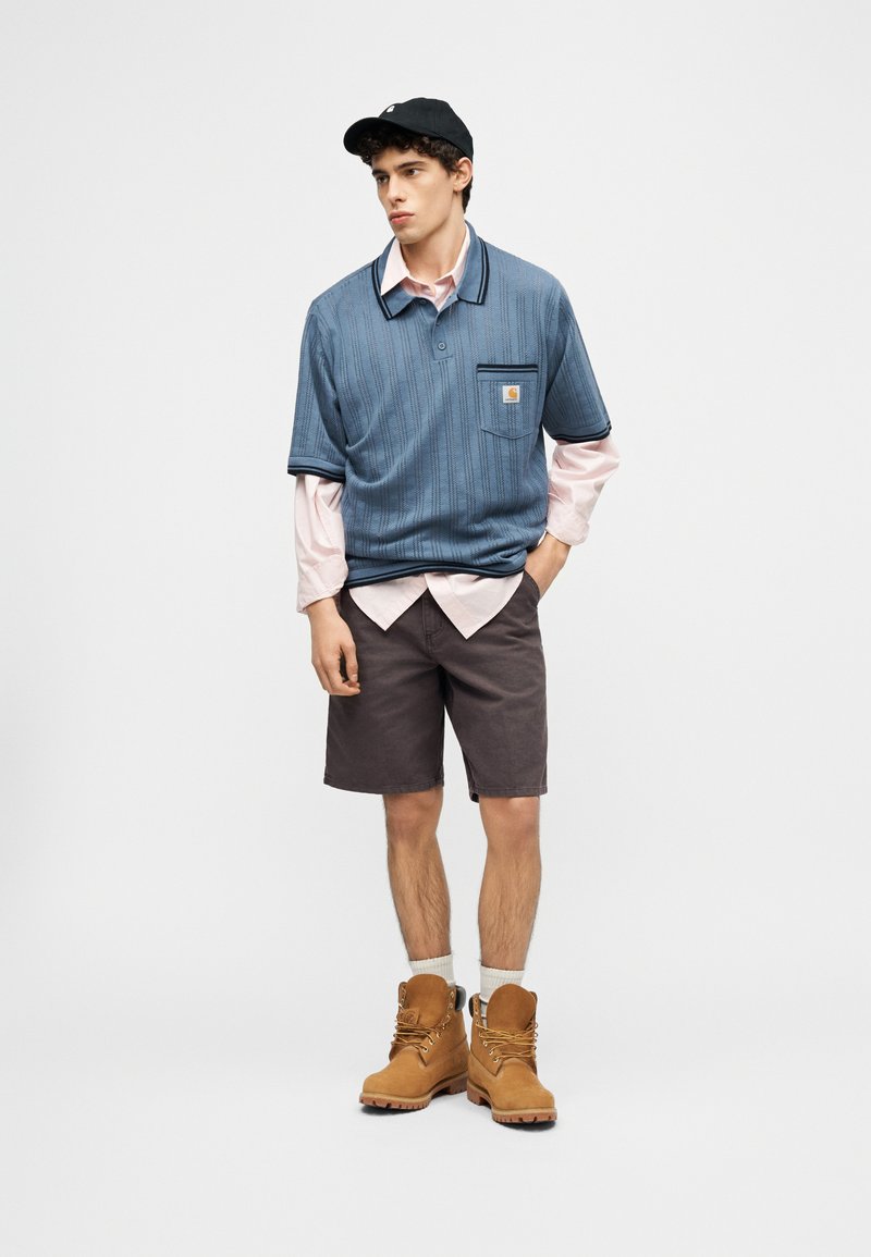 Jonge man met een zwarte pet, een blauw shirt met korte mouwen over een roze shirt met lange mouwen, donkere korte broek, witte sokken en beige laarzen, staand met één hand in zijn broekzak.