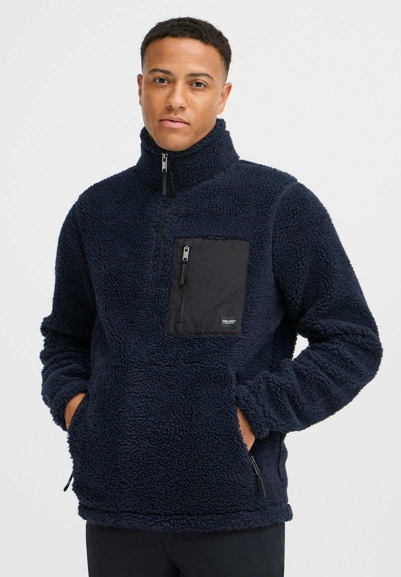 Navy fleece pullover with high collar and zippered front pocket; features textured fabric and side zip pockets for added utility.