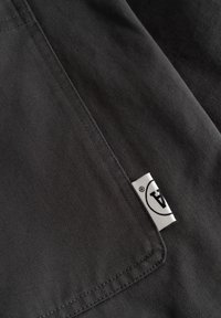 Gros plan sur un tissu sombre avec des coutures détaillées et une petite étiquette blanche portant un logo circulaire noir avec des lettres à l'intérieur.