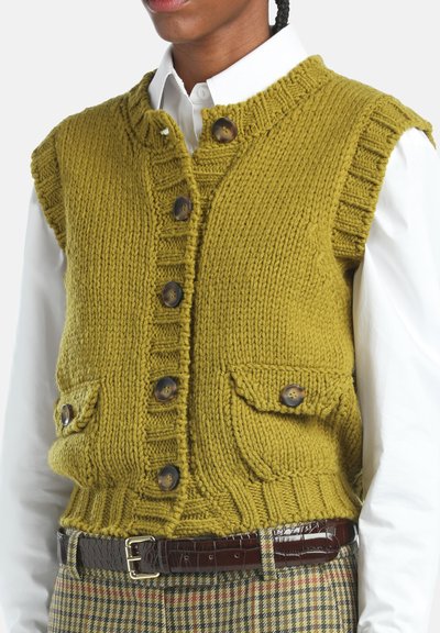 Personne portant un cardigan sans manches en laine jaune moutarde avec des boutons et des poches à rabat sur une chemise blanche et un pantalon à carreaux avec une ceinture brune.