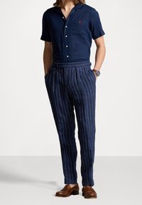 Navy short-sleeve linen button-up shirt with a small red logo, paired with pinstriped navy trousers and brown leather shoes.