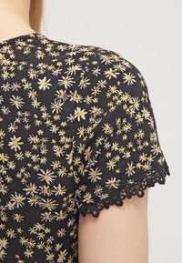 Czarna bluzka z materiału z printem stokrotek w kolorze białym i żółtym, z falbaniastym koronkowym wykończeniem przy krawędziach rękawów.