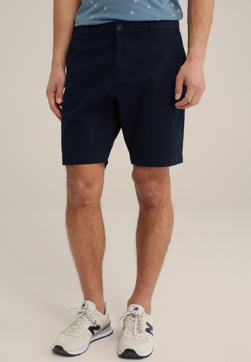 Shorts en coton bleu marine avec fermeture à bouton, coupe droite et poches latérales. Portés avec des baskets crème avec des accents bleu marine.