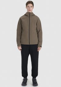 Jeune homme debout face à l’avant, portant une veste à capuche marron zippée, un pantalon noir et des baskets noires, sur un fond clair et uni.