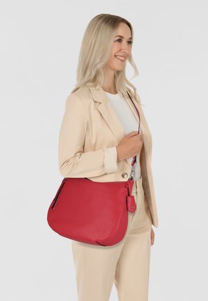 Sac à bandoulière en cuir rouge avec une forme courbée et une texture lisse, doté d'une bandoulière amovible et d'une petite étiquette sur le côté.