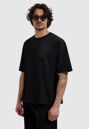 Man with curly hair and sunglasses wearing black oversized t-shirt and pants, showing tattoos on right forearm and upper arm.