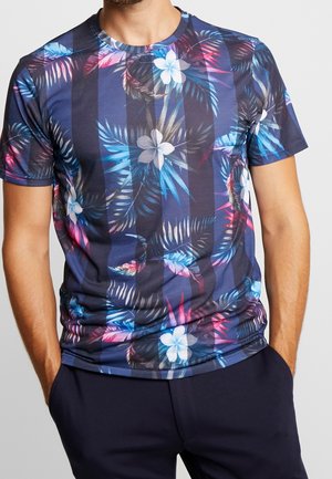Mężczyzna w granatowej koszulce z krótkim rękawem, z pionowymi paskami i żywym tropikalnym kwiatowym nadrukiem, z rękami w kieszeniach ciemnych spodni, na szarym tle.