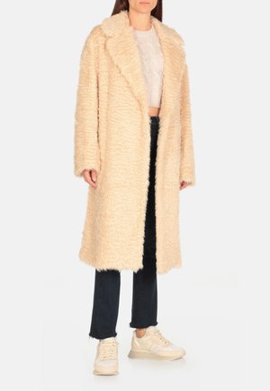 Manteau en fausse fourrure beige clair avec un col cranté, des manches longues et une coupe décontractée. Porté sur un haut en tricot et un jean noir, associé à des baskets.