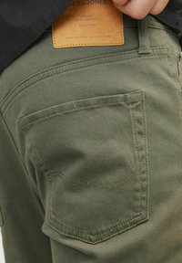 Pantaloni verde oliva con tessuto testurizzato, dotati di una tasca posteriore, dettagli di cucitura visibili e un'etichetta in pelle con informazioni sul marchio.