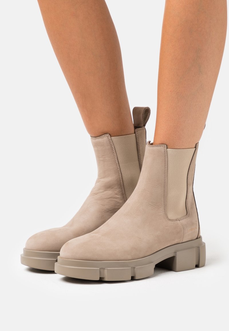 Bottes en cuir beige montant à la cheville avec semelles épaisses portées sur des jambes nues, dotées de panneaux élastiques latéraux et de languettes à l'arrière.
