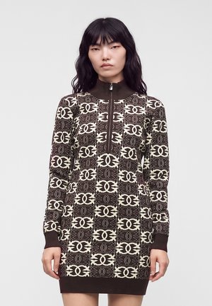 Femme aux longs cheveux noirs portant une robe en maille marron et crème à motifs, manches longues, avec une fermeture éclair devant et un col montant, face à l'avant.