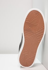 Baskets noires avec une semelle en caoutchouc brun texturé et une semelle intermédiaire blanche, présentant des motifs géométriques pour l'adhérence et la stabilité.