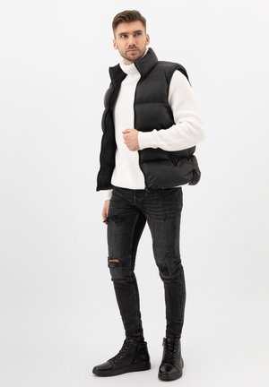 Gilet imbottito nero sopra un dolcevita bianco, con jeans neri strappati e sneakers alte nere. Texture lisce e design a strati.