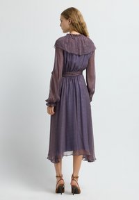 Lang, vloeiende jurk in paarse en rode paisleyprint, met een gerimpelde kraag, lange mouwen en een cinched taille. Gecombineerd met bruin gekleurde sandalen met bandjes.