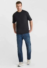 Camiseta negra de manga corta combinada con vaqueros azules que presentan múltiples bolsillos y costuras. Zapatillas deportivas en gris claro con detalles reflectantes.