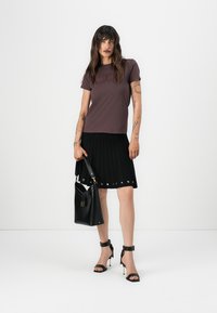 T-shirt marron ajusté, jupe noire plissée avec détails en œillets, sac à main noir et sandales noires à talons avec des accents dorés.