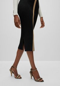 Jupe en maille côtelée noire avec une bande latérale dorée, longueur au genou, dotée d'une fente sur le côté. Associée à des sandales métalliques à talons hauts et à lanières.