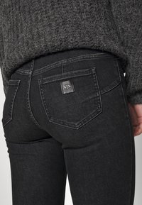 Jeans de mezclilla negra con un diseño ajustado, que presentan un logotipo distintivo en el bolsillo trasero y una textura ligeramente tejida.