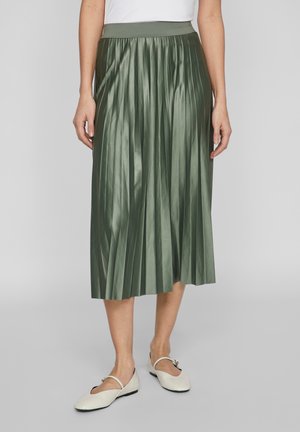 Kvinde iført en højtaljet grøn metallic plisseret midi-nederdel kombineret med hvide Mary Jane-flats mod en ensfarvet baggrund.