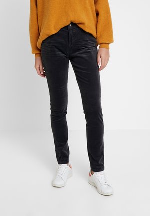 Personne portant un pantalon noir slim, des baskets blanches avec des accents orange et un pull ample orange, debout sur un sol blanc avec un fond uni.