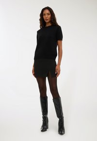 Kobieta w czarnej krótkiej bluzie, czarnej mini spódniczce z przeszyciem z przodu, przejrzystych czarnych rajstopach oraz wysokich czarnych skórzanych kozakach.
