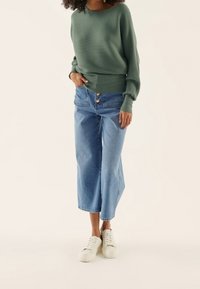 Grüner, gerippter Sweatshirt mit weiten Ärmeln, kombiniert mit hellblauen, cropped Jeans mit zwei Knöpfen in der hohen Taille und weißen Turnschuhen.