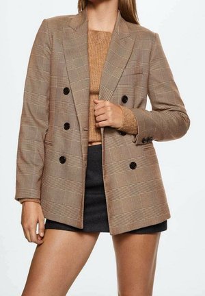 Femme portant un blazer marron à carreaux croisé sur un pull en tricot beige et un short noir sur un fond blanc uni.