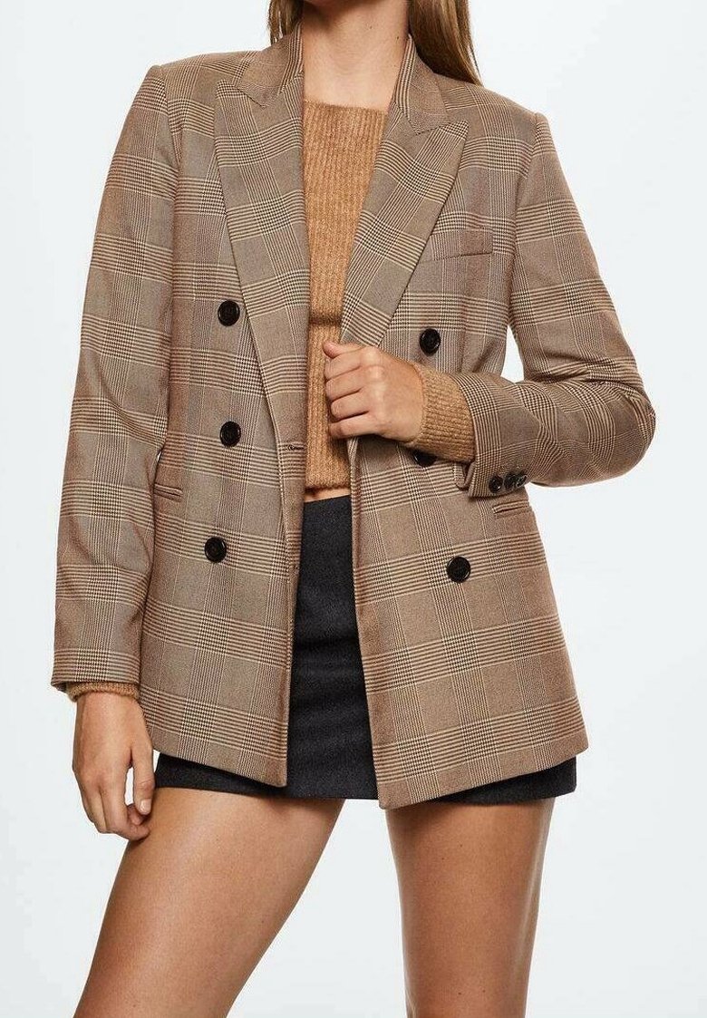 Femme portant un blazer marron à carreaux croisé sur un pull en tricot beige et un short noir sur un fond blanc uni.