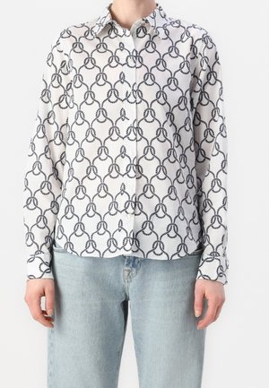 Personne portant une chemise blanche à manches longues avec un motif de boucles noires reliées et un jean bleu clair, debout devant un fond uni.