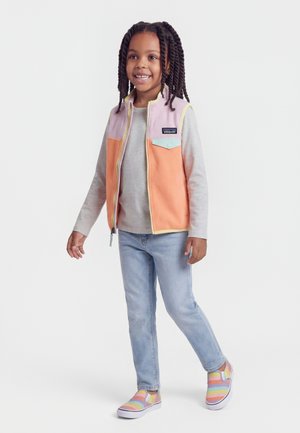 Χαμογελαστό παιδί με πλεξούδες, ντυμένο με γιλέκο σε διαφορετικά χρώματα, γκρι μπλούζα με μακριά μανίκια, ανοιχτόχρωμο τζιν και ριγέ παπούτσια slip-on, στέκεται σε λευκό φόντο.