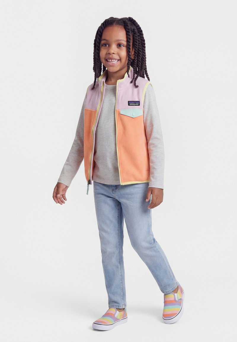 Χαμογελαστό παιδί με πλεξούδες, ντυμένο με γιλέκο σε διαφορετικά χρώματα, γκρι μπλούζα με μακριά μανίκια, ανοιχτόχρωμο τζιν και ριγέ παπούτσια slip-on, στέκεται σε λευκό φόντο.