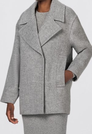 Personne portant un manteau en laine gris clair à larges revers et poches latérales, par-dessus une robe en maille grise assortie, une main dans la poche.