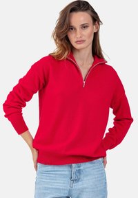 Pull rouge à col zippé en tissu tricot doux. Comporte des manches longues et des poignets côtelés, associé à un jean bleu clair.