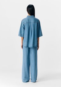 Femme aux longs cheveux bruns portant une chemise en jean bleu clair ample et un jean à jambes larges, debout de dos sur un fond blanc.