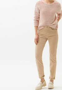 Pull en tricot rose clair à texture lâche, associé à un pantalon beige ajusté ; la personne porte des chaussures plates crème, debout de manière décontractée.