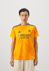 Žuta atletska majica s okruglim izrezom, crnim detaljima i uzorkom dijagonalnih linija. Na majici piše "Emirates FLY BETTER" i prisutan je logo.