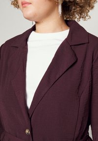 Blazer burdeos con una tela texturizada, con cuello de solapa y botón metálico, superpuesto sobre una camisa blanca.
